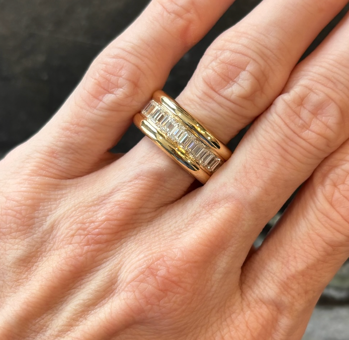 A close-up of a hand wearing two rings on the ring finger: one is plain 18K yellow gold, and the other is the Channel Set Emerald Cut Diamond Eternity Band featuring a row of rectangular emerald cut diamonds set horizontally.