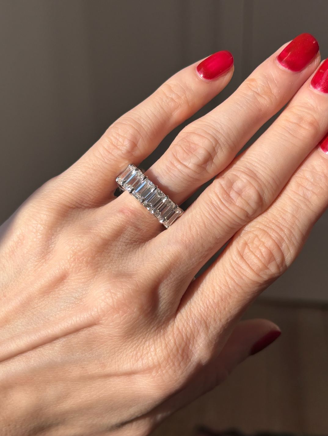 A hand with red nails wears the Emerald Cut Diamond Eternity Band, a silver ring adorned with rectangular emerald-cut diamonds on the ring finger against a softly lit, blurred background.