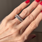 A hand with red nails wears the Emerald Cut Diamond Eternity Band, a silver ring adorned with rectangular emerald-cut diamonds on the ring finger against a softly lit, blurred background.