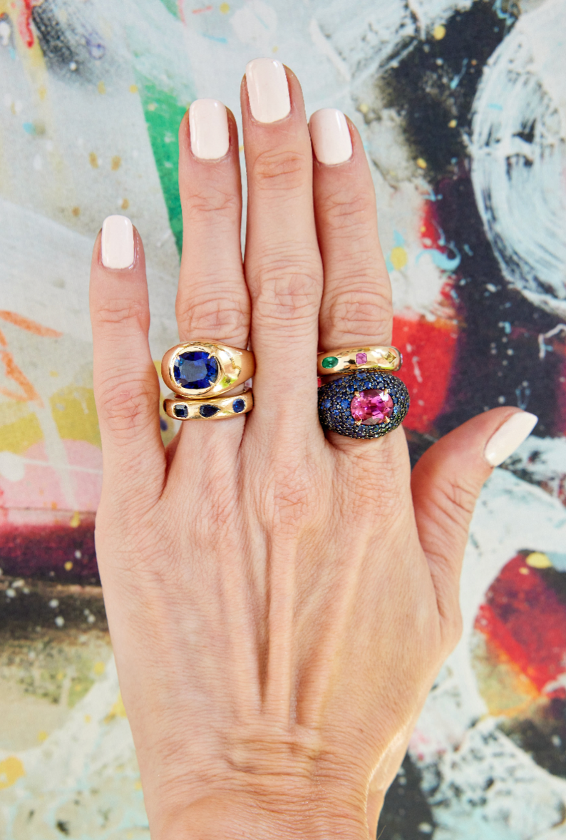 A hand with light skin and pale pink nails models four gemstone rings, including the Oval Gemstone Bombé Ring in Yellow Gold, on the index and middle fingers, set against a vibrant abstract multicolored background.