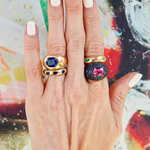 A hand with light skin and pale pink nails models four gemstone rings, including the Oval Gemstone Bombé Ring in Yellow Gold, on the index and middle fingers, set against a vibrant abstract multicolored background.