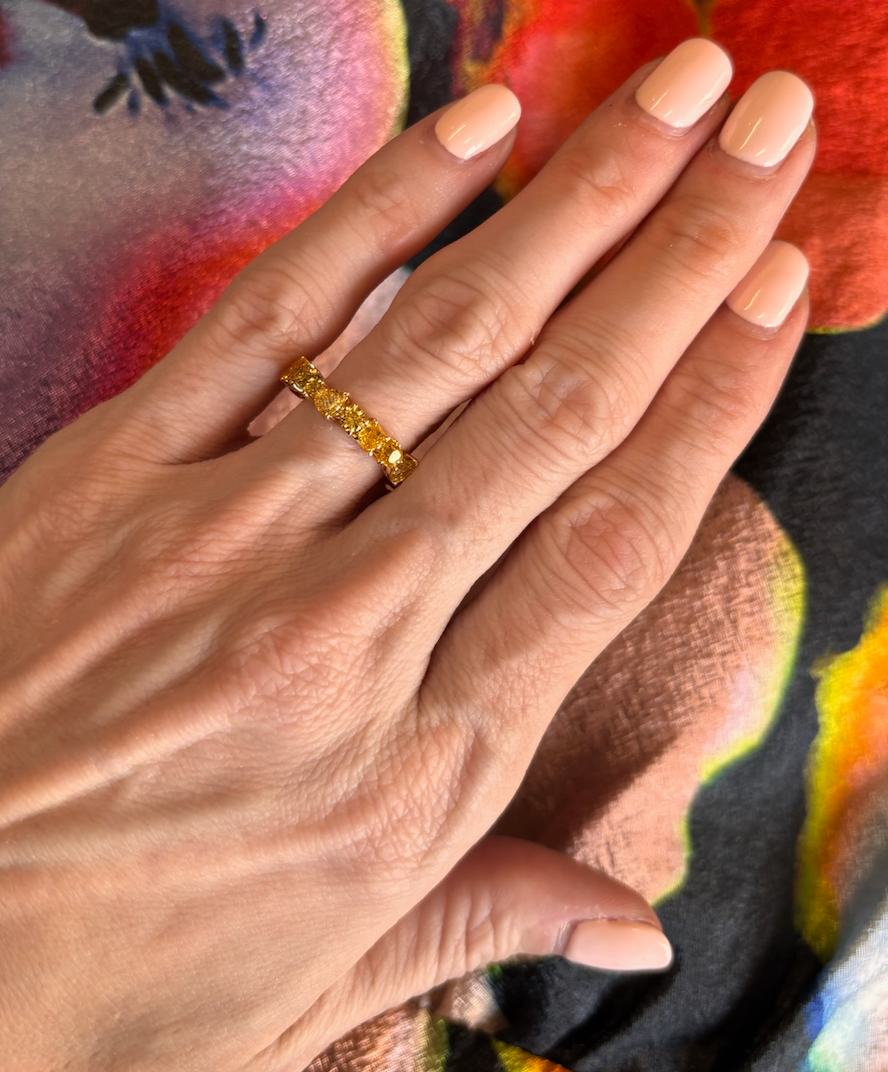A hand with pale pink manicured nails showcases the Mixed Shape Eternity Band in Yellow Gold with Yellow Diamonds, set against a vibrant, multicolored fabric background.