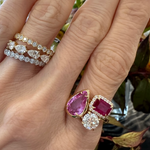 A hand wearing three 18K yellow gold diamond rings on the index finger and a Cluster Ring in yellow gold with bezel-set pear shape, pavé emerald cut sapphires, and round diamond on the middle finger. Green and brown leaves in the background.