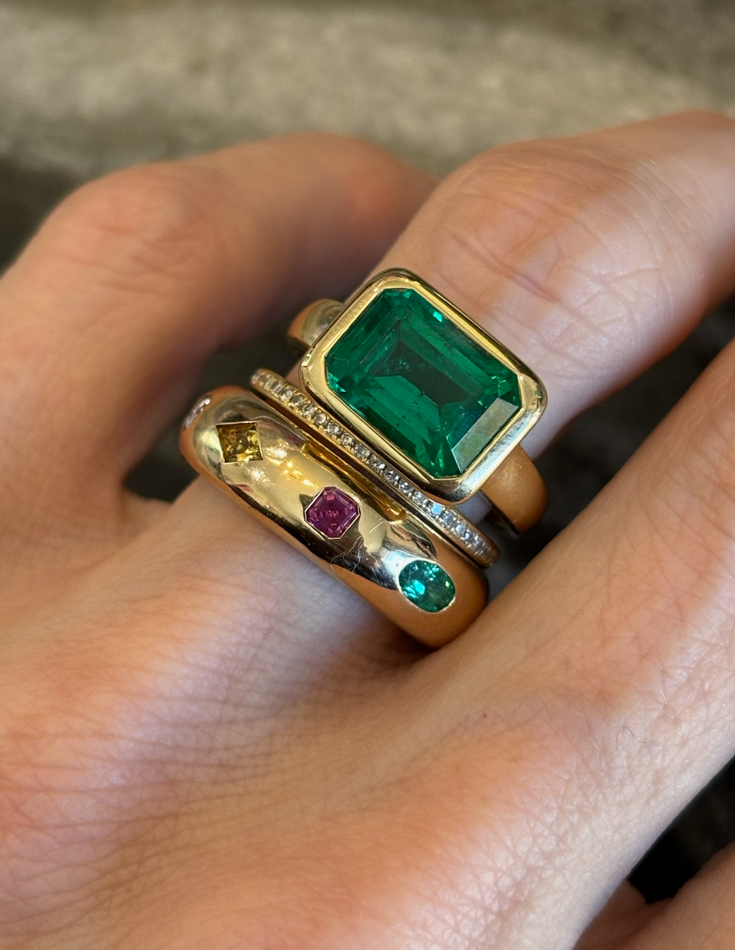 A hand wearing three gold rings: the East-West Bezel Set Emerald Cut Emerald Engagement Ring in yellow gold, a thin diamond band, and a wide 18K yellow gold band with green, pink, and yellow gemstones.