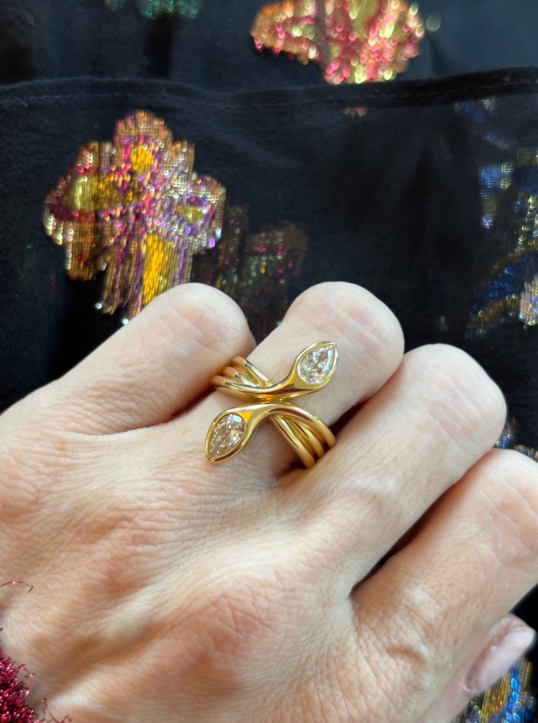 A hand wearing the Double Headed Serpent Ring with Pear Diamonds in 18k yellow gold—featuring two crossing bands ending in pear-shaped diamonds—holds dark fabric embroidered with colorful flowers.