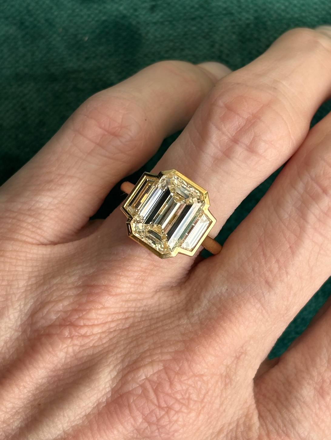 A close-up of a hand wearing the Emerald Cut Diamond Engagement Ring with Bezel Setting and Trapezoid Side Stones, set against textured green fabric for an elegant, luxurious effect.