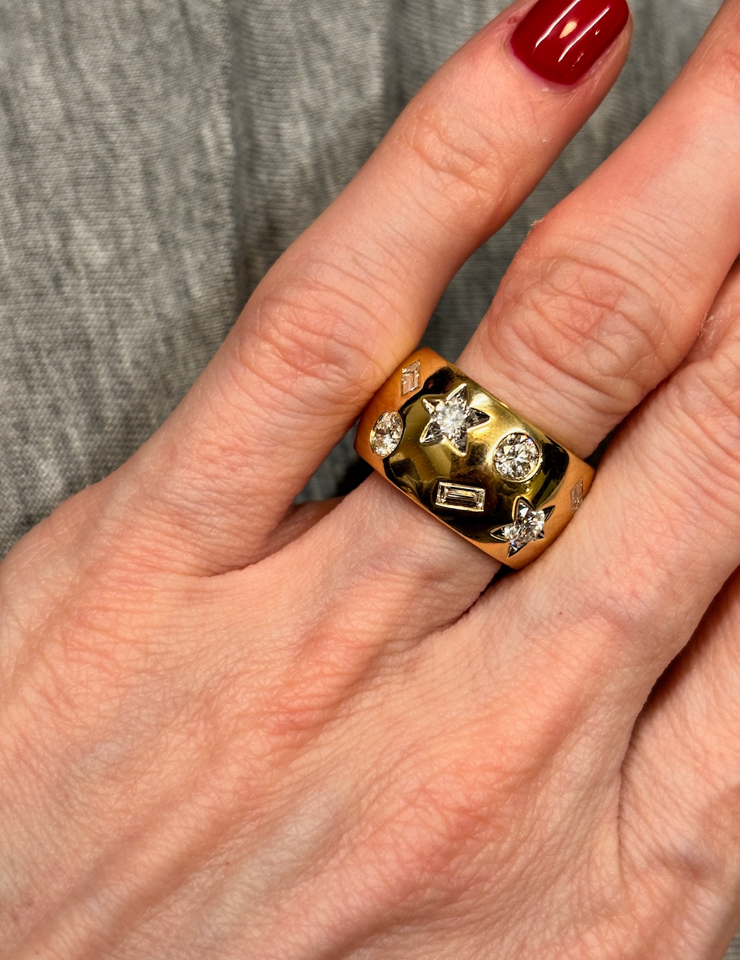 A hand with red-painted nails wears the Large Celestial Cigar Band in Yellow Gold with mixed shape diamonds, set against a gray fabric background.