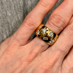 A hand with red-painted nails wears the Large Celestial Cigar Band in Yellow Gold with mixed shape diamonds, set against a gray fabric background.