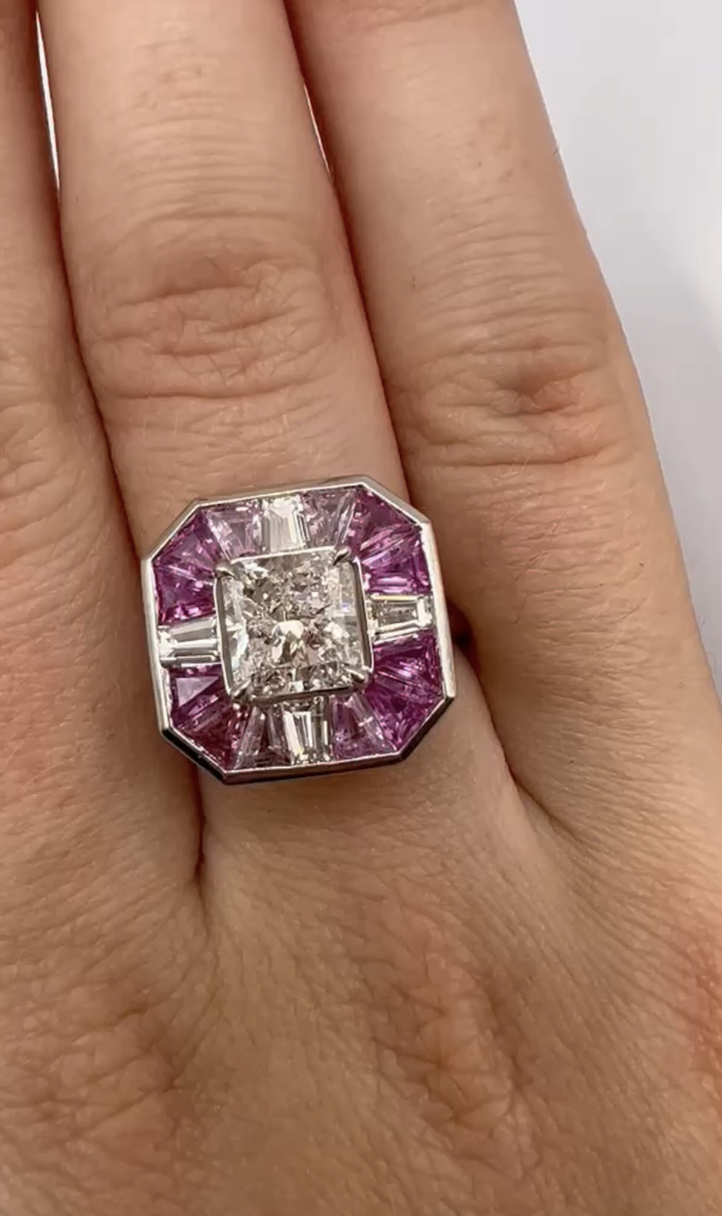The Mosaic Ring features a radiant cut diamond at the center, surrounded by pink sapphires and baguette diamonds in a square design, worn on the finger.