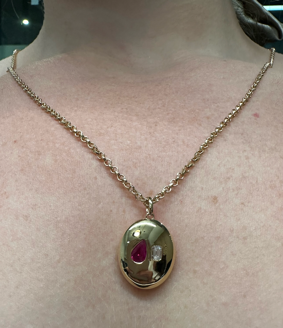The Bespoke Burnish Set Pear Shape Ruby and Emerald Cut Diamond in Yellow Gold is a pendant necklace featuring a round yellow gold charm with a red pear-shaped ruby and a small clear emerald cut diamond.