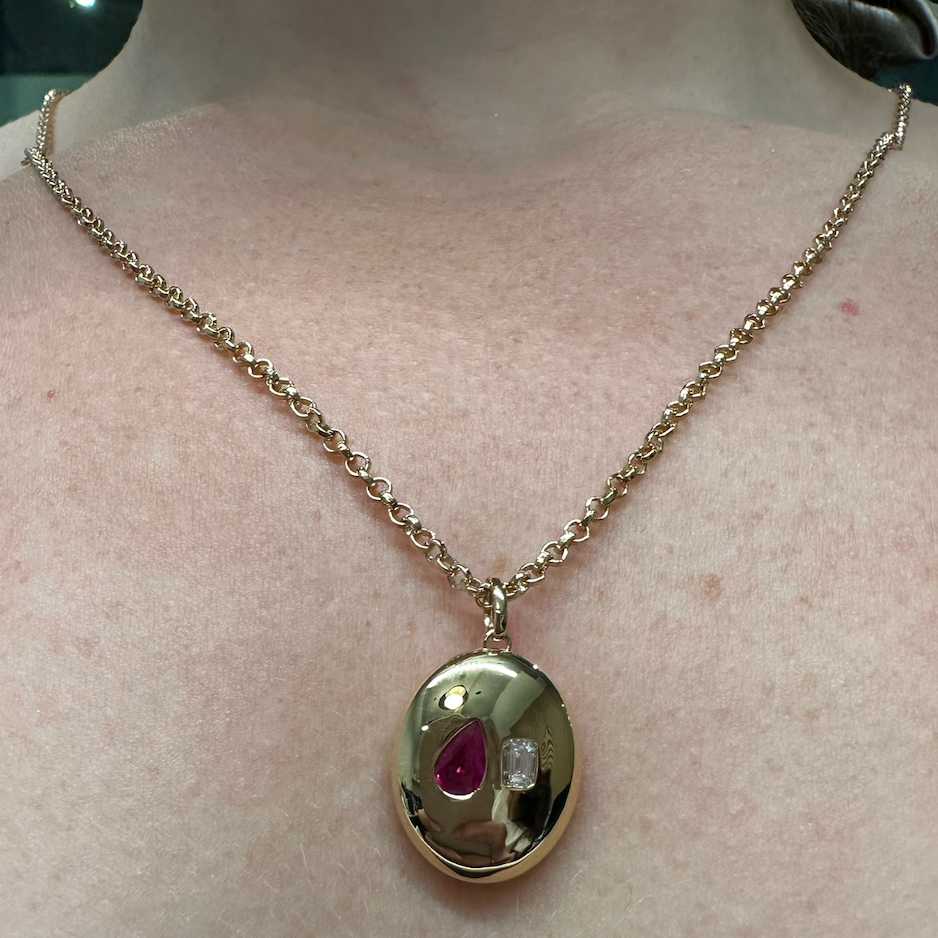 The Bespoke Burnish Set Pear Shape Ruby and Emerald Cut Diamond in Yellow Gold is a pendant necklace featuring a round yellow gold charm with a red pear-shaped ruby and a small clear emerald cut diamond.