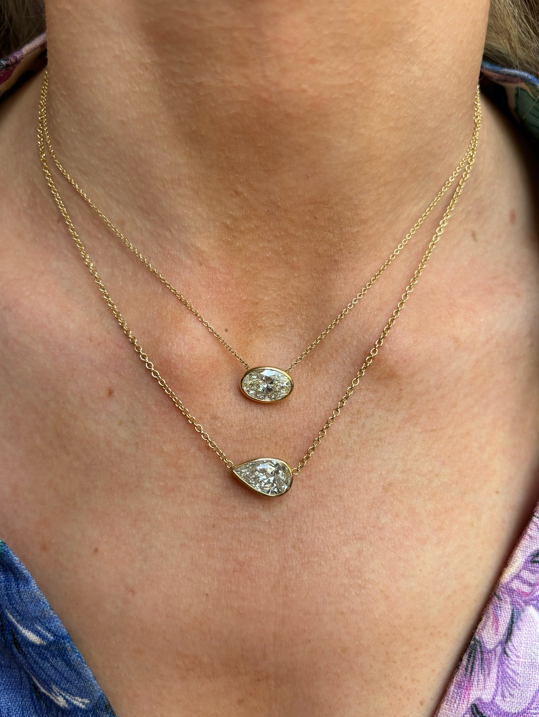 A close-up of a neck adorned with two delicate 18k yellow gold necklaces, including the East-West Bezel Set Oval Shape Diamond Pendant Necklace. The person wears a floral-patterned blue and purple garment.