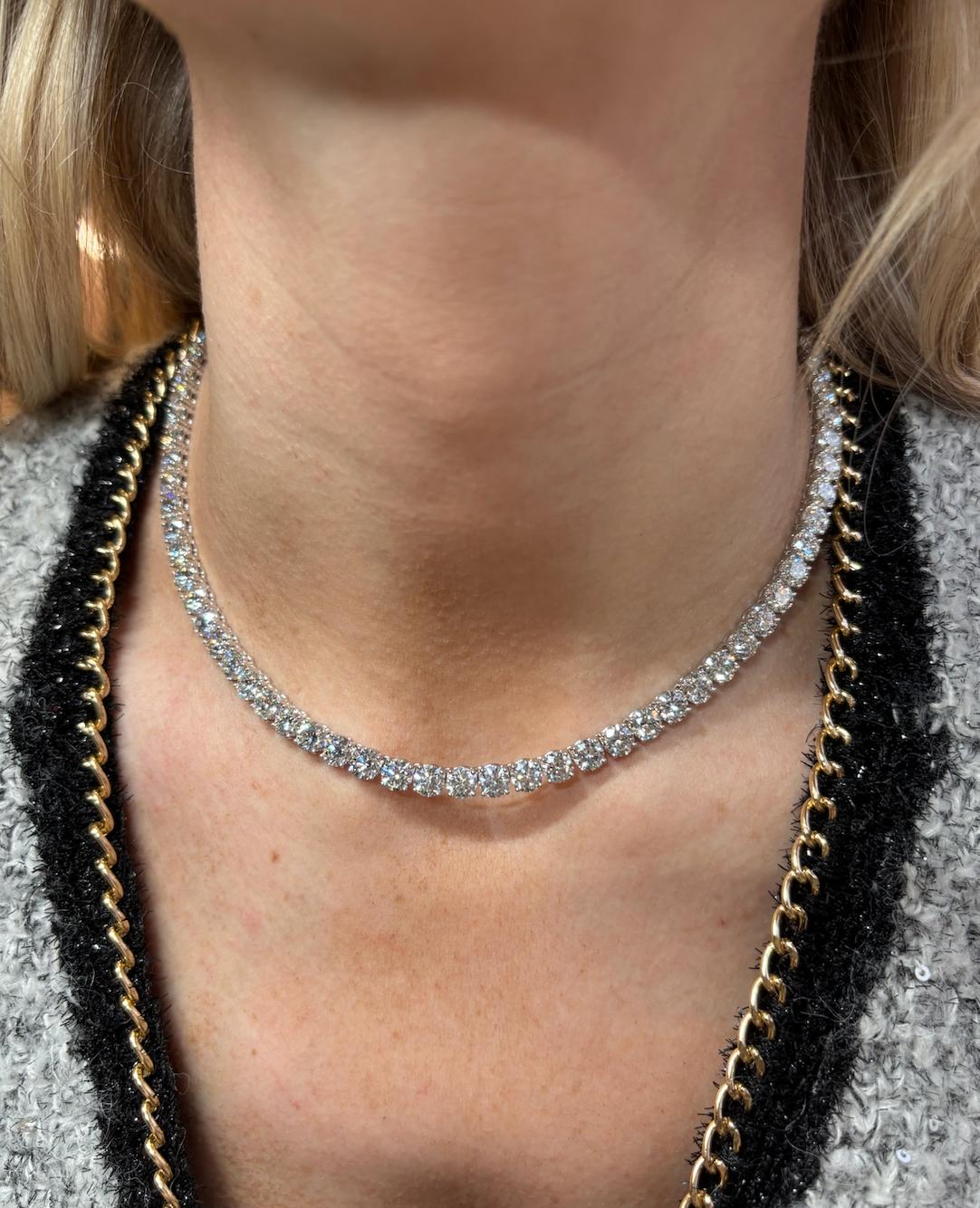 A close-up of a person's neck wearing a Large Round Diamond Tennis Necklace, paired with a gray textured garment featuring 18K gold and black chain trim.