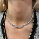 A close-up of a person's neck wearing a Large Round Diamond Tennis Necklace, paired with a gray textured garment featuring 18K gold and black chain trim.