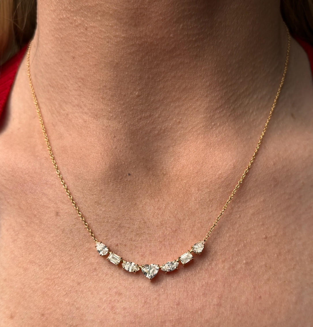 A close-up of the Mixed Shape Diamond Pendant Necklace, crafted in 18K gold, showcases seven clear diamonds—including a Heart Shape Diamond—set along its delicate chain and worn gracefully around the neck.