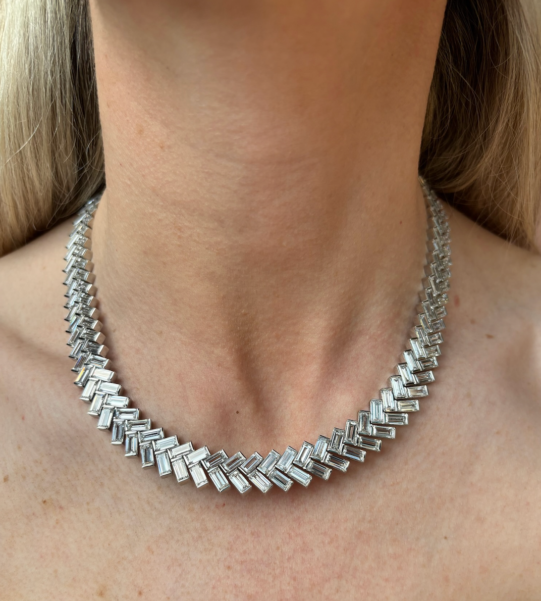 A close-up of a woman's neck showcases the Bezel Set Chevron Diamond Tennis Necklace in White Gold, featuring a geometric chevron pattern with baguette diamonds and rectangular links. The blurred background emphasizes its intricate design.