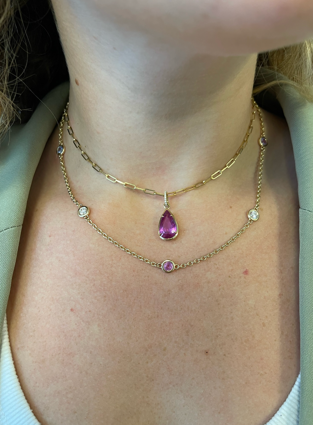 A person wearing a layered 18K yellow gold necklace, including the Station Necklace in Yellow Gold with Round Diamonds and Multicolor Sapphires, over a white top.