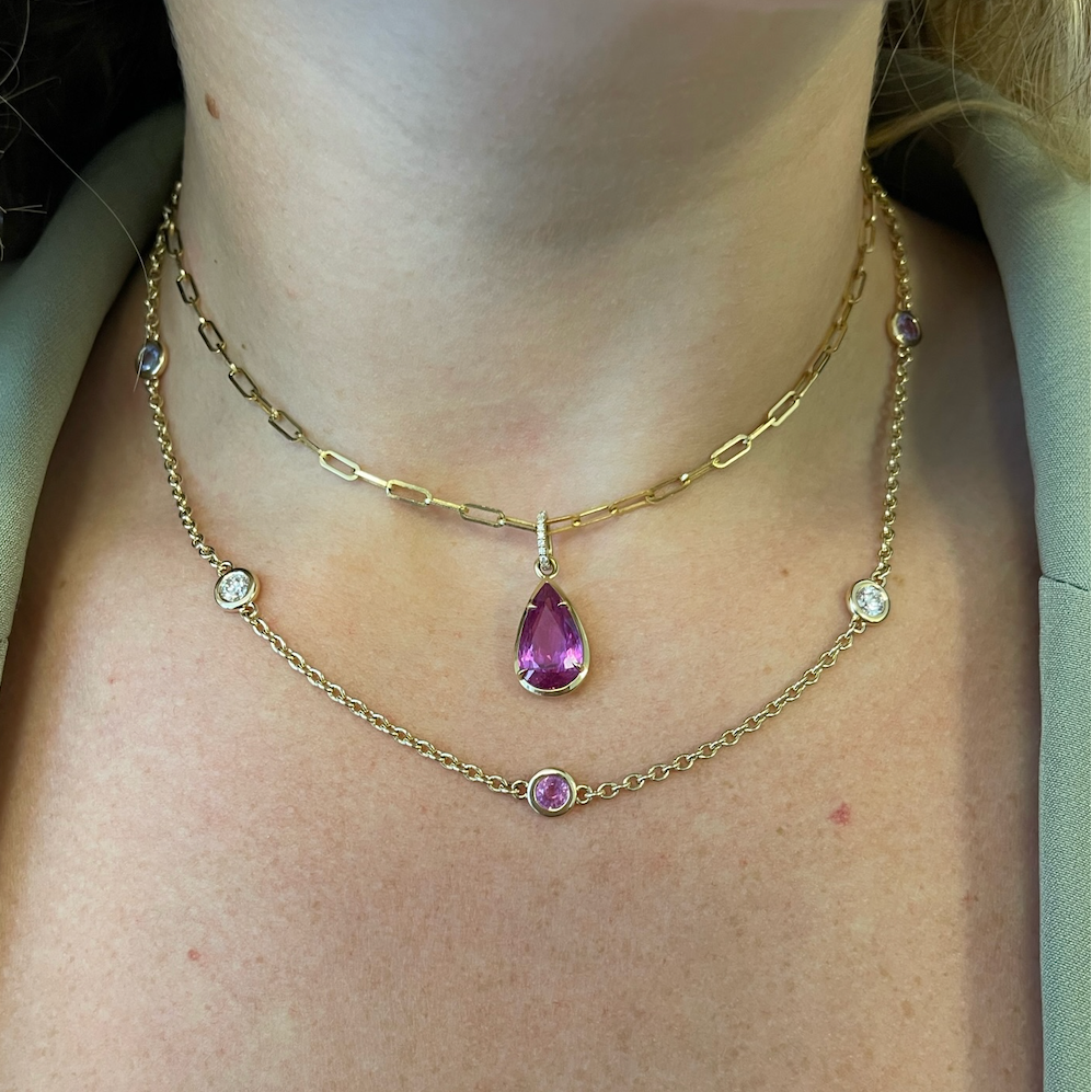 A person wearing a layered 18K yellow gold necklace, including the Station Necklace in Yellow Gold with Round Diamonds and Multicolor Sapphires, over a white top.