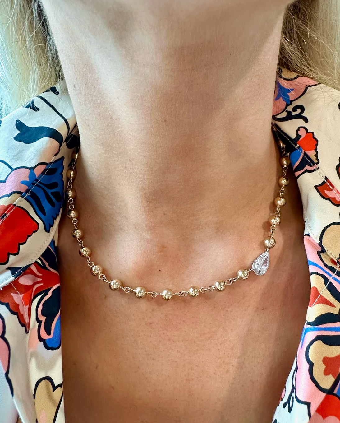 A close-up of a woman's neck wearing the Garland Chain Necklace in Yellow Gold, featuring an east-west set pear diamond. She pairs it with a vibrant blouse adorned with floral and abstract patterns.