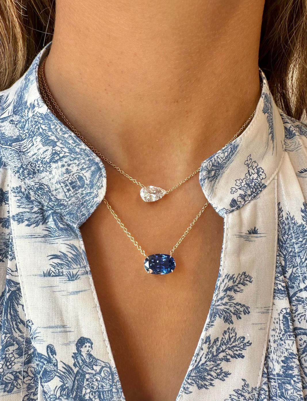 A close-up of a person wearing a blue and white patterned blouse and two gold chain necklaces, one in 18K yellow gold with a clear teardrop pendant and the other featuring the East-West Oval Sapphire Pendant in yellow gold.