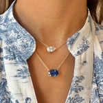 A close-up of a person wearing a blue and white patterned blouse and two gold chain necklaces, one in 18K yellow gold with a clear teardrop pendant and the other featuring the East-West Oval Sapphire Pendant in yellow gold.