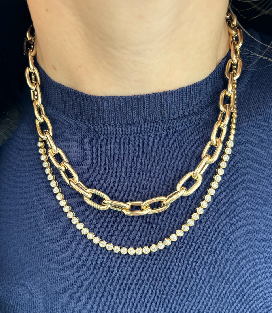 A person wears a blue top with two gold necklaces: the Serpentine Signature Chain Necklace, an 18-inch 14K yellow gold chain with large links, and another adorned with small, round, sparkling stones.