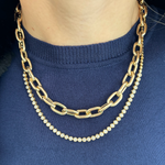 A person wears a blue top with two gold necklaces: the Serpentine Signature Chain Necklace, an 18-inch 14K yellow gold chain with large links, and another adorned with small, round, sparkling stones.