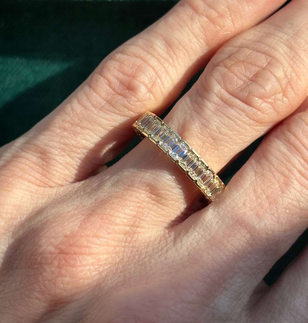 A close-up of a hand wearing the Half Bezel Emerald Cut Diamond Eternity Band on the middle finger, its rectangular diamonds reflecting light against a dark green background.