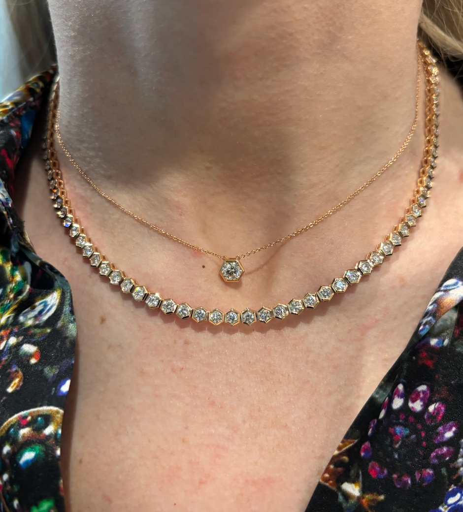 A close-up of a person wearing two necklaces: the Hexagon Bezel Set Round Brilliant Diamond Pendant and a solitaire necklace with round, sparkling stones, styled over a colorful, patterned top.