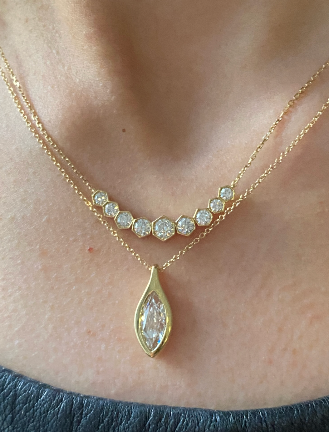 A close-up of a person wearing the Serpent Bezel Set Marquise Diamond Solitaire Pendant in 18k gold, layered with another necklace featuring hexagon-shaped bezel set diamonds, both resting on bare skin above a dark top.