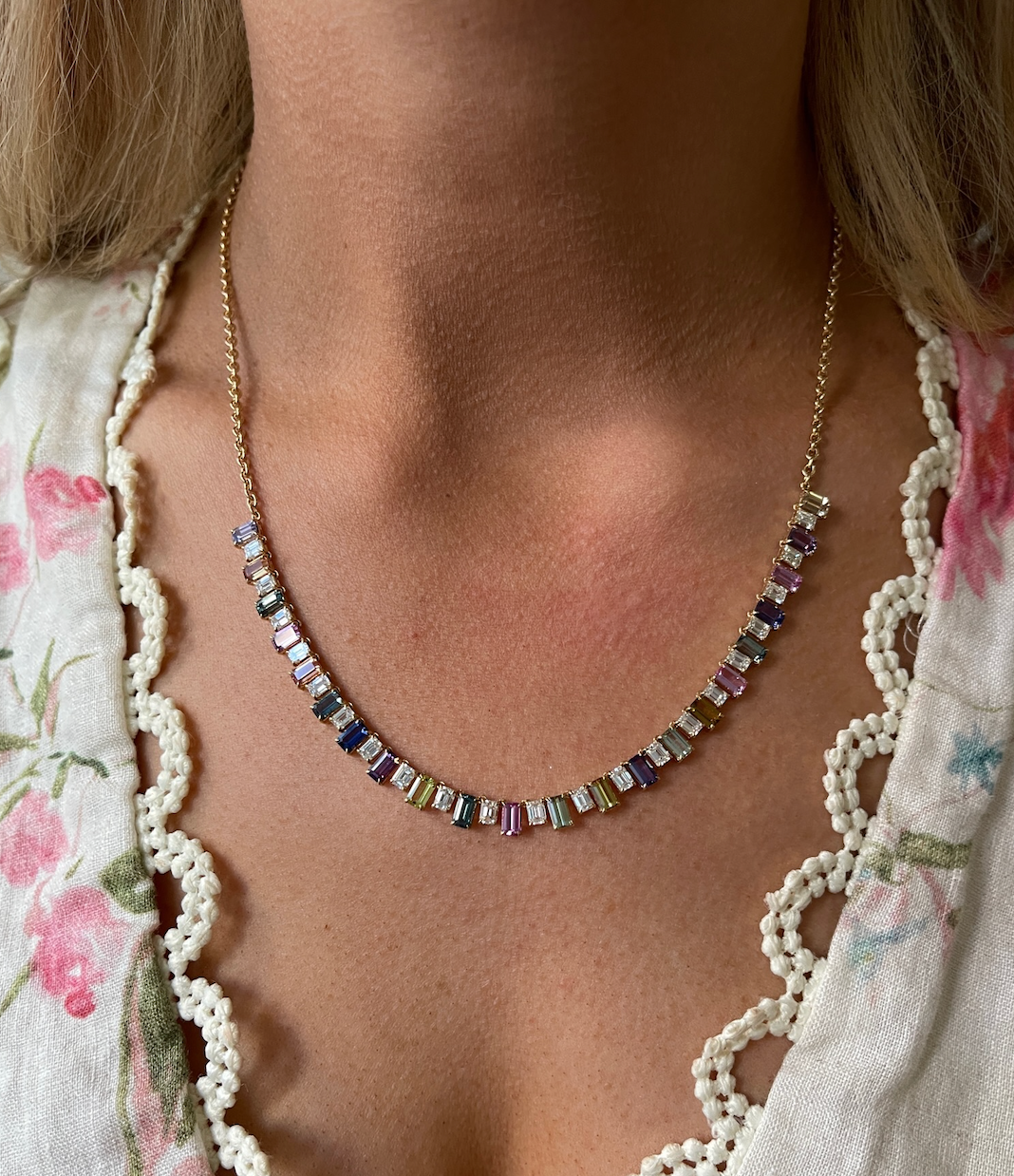 A close-up of a person wearing the Confetti Necklace in Yellow Gold with Multicolor Emerald Cut Sapphires, paired with a floral-patterned blouse featuring a scalloped lace trim.