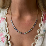 A close-up of a person wearing the Confetti Necklace in Yellow Gold with Multicolor Emerald Cut Sapphires, paired with a floral-patterned blouse featuring a scalloped lace trim.