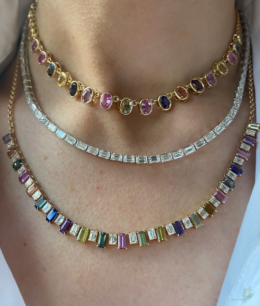 A close-up of a person’s neck wearing three layered necklaces: a gemstone choker, a bezel set tennis necklace, and the Gumdrop Necklace in Yellow Gold with Oval Multicolor Sapphires.