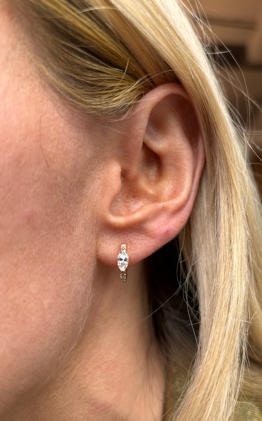 Close-up of a woman's ear with straight blonde hair, wearing Pavé Huggies with Marquise Shape Diamond in 18k gold, accented by a marquise-shaped center stone and two smaller round diamonds above it.