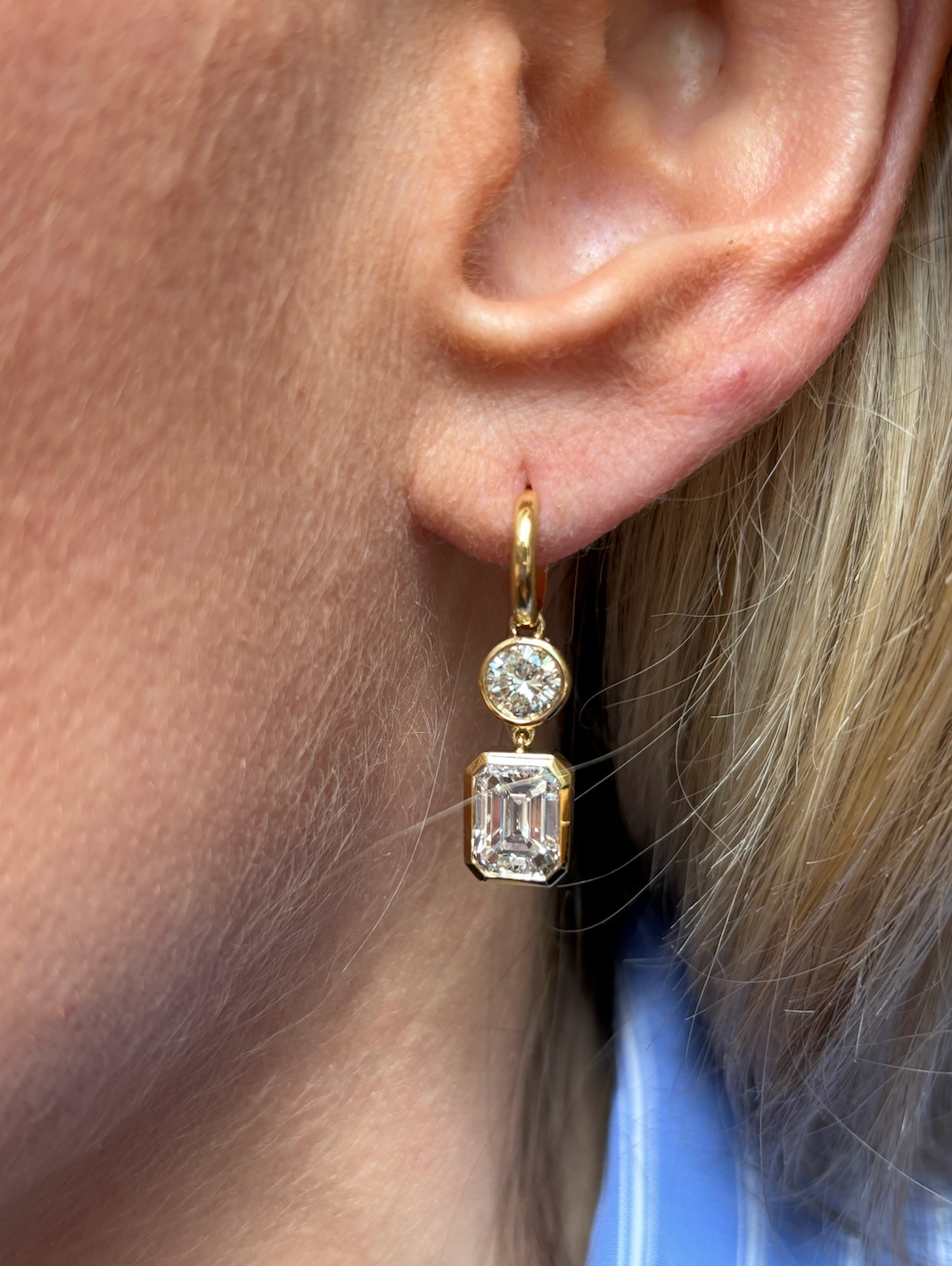 A person with blonde hair and a blue and white striped shirt wears Asymmetrical Drop Earrings with Bezel Set Round and Emerald Cut Diamonds in 18K yellow gold.