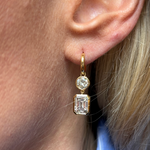 A person with blonde hair and a blue and white striped shirt wears Asymmetrical Drop Earrings with Bezel Set Round and Emerald Cut Diamonds in 18K yellow gold.