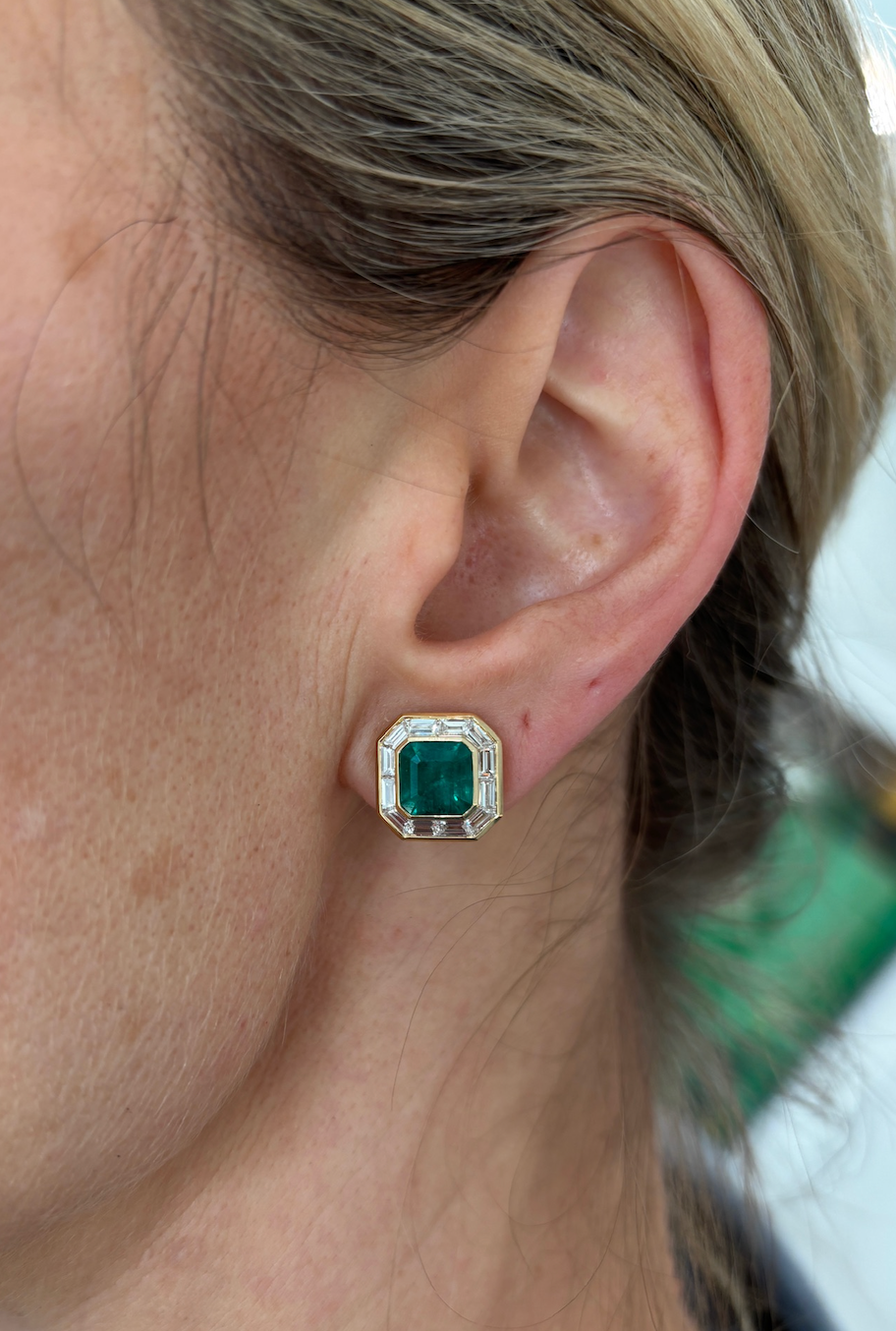 Close-up of a light-skinned person with blonde hair wearing the Mosaic Studs in Yellow Gold, featuring Asscher Cut Emeralds and baguette diamonds in a geometric setting.
