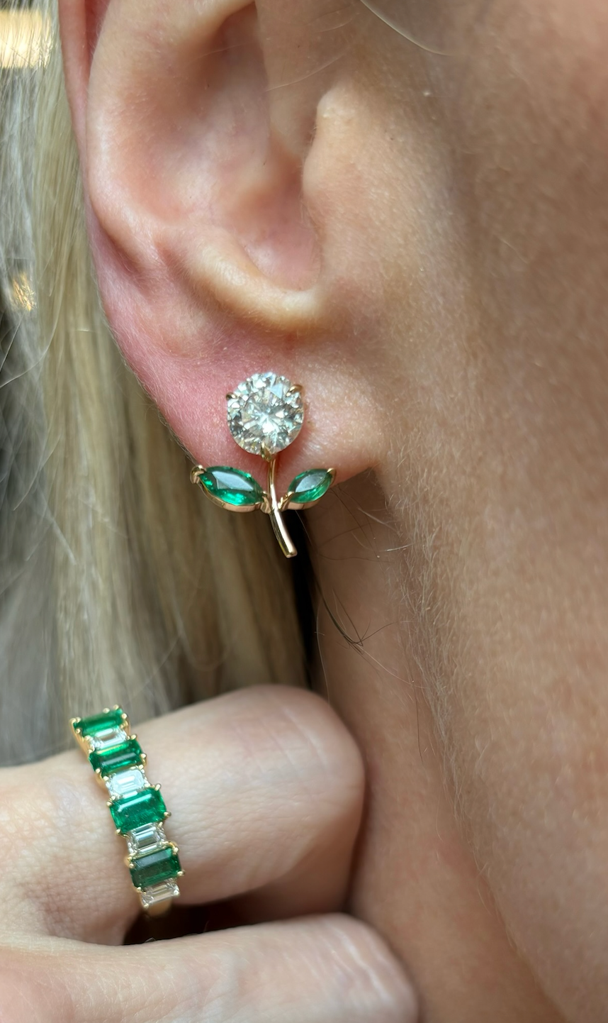 A close-up of an ear wearing Flower Ear Jackets with marquise emerald petals in yellow gold. A hand with an 18K yellow gold ring set with three rectangular green stones is also visible.