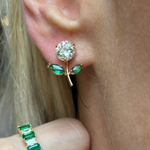 A close-up of an ear wearing Flower Ear Jackets with marquise emerald petals in yellow gold. A hand with an 18K yellow gold ring set with three rectangular green stones is also visible.