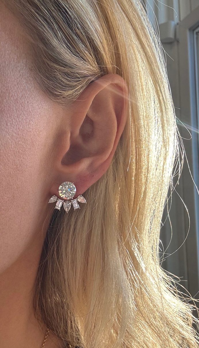 Close-up of a person's ear wearing Alternating Pear Shape and Emerald Cut Diamond Ear Jackets in 18K white gold, with sunlight catching their blonde hair.