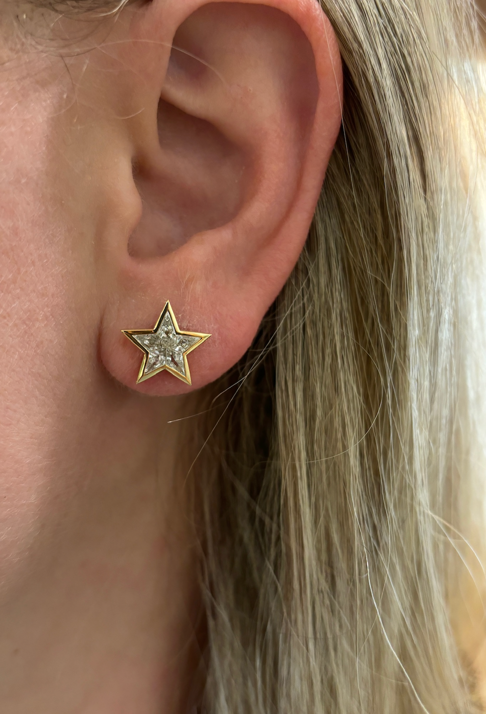 A close-up of an ear adorned with Bezel Set Kite Diamond Star Studs in yellow gold, showcasing a central kite diamond. Blonde hair is partially visible on the right side of the image.