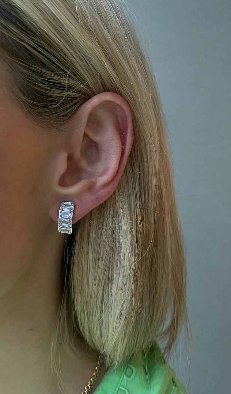 Close-up of a left ear with short blonde hair wearing the J Hoop Earring in white gold, featuring bracket bezel set emerald cut diamonds. The person wears a light green top; the earring's design echoes an emerald cut diamond style.