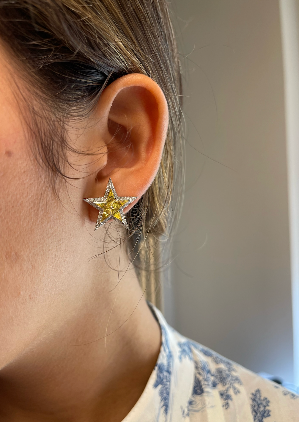 Close-up of a person’s ear adorned with Celestial Star Studs in white gold, featuring diamond pavé and yellow kite sapphires. The wearer has brown hair and is dressed in a white top with blue floral patterns.