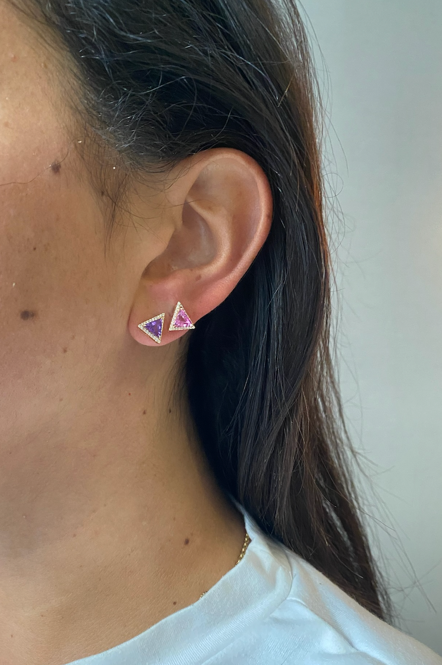 Close-up of a person with long brown hair wearing a white top and two This & That Pavé Trinity Studs featuring multicolor trillion cut sapphires—one purple, one pink—in 18K yellow gold.