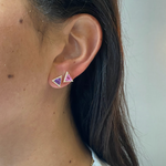 Close-up of a person with long brown hair wearing a white top and two This & That Pavé Trinity Studs featuring multicolor trillion cut sapphires—one purple, one pink—in 18K yellow gold.