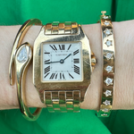 A wrist adorned with a gold Cartier watch, the Serpent Pear Diamond Bangle in 18K yellow gold, and a pear-shaped diamond bracelet, set against a green background.