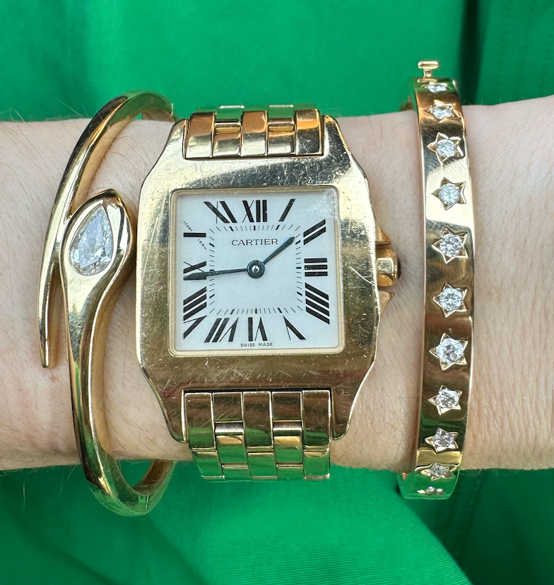 A wrist adorned with a gold Cartier watch, the Serpent Pear Diamond Bangle in 18K yellow gold, and a pear-shaped diamond bracelet, set against a green background.
