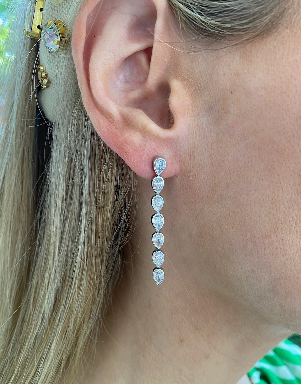 Close-up of a woman wearing Chasing Pears Drop Earrings in white gold, bezel set with seven pear diamonds in a vertical line. Her straight blonde hair is tucked behind her ear and accented with a decorative hairpin.