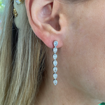 Close-up of a woman wearing Chasing Pears Drop Earrings in white gold, bezel set with seven pear diamonds in a vertical line. Her straight blonde hair is tucked behind her ear and accented with a decorative hairpin.