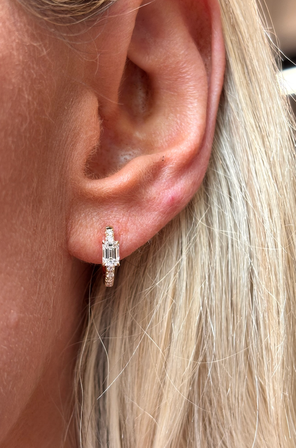 A close-up of a person's ear with light blonde hair, wearing the Pavé Huggie with Emerald Cut Diamonds, a gold hoop earring featuring emerald cut diamonds and rectangular clear gemstones.